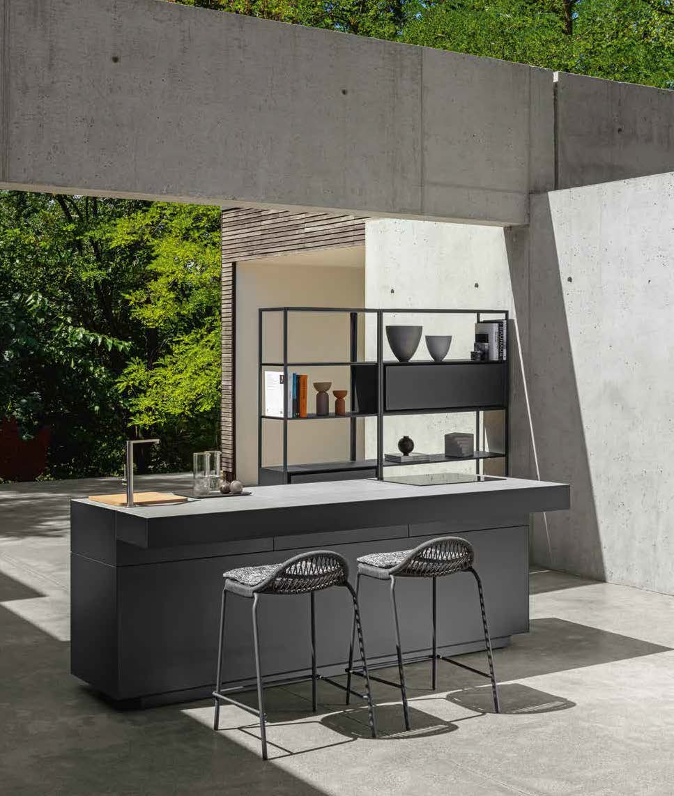 Cuisine extérieure moderne comprenant un îlot minimaliste, deux tabourets de bar tressés, des étagères ouvertes avec des objets décoratifs et des murs en béton entourés d'une végétation luxuriante.