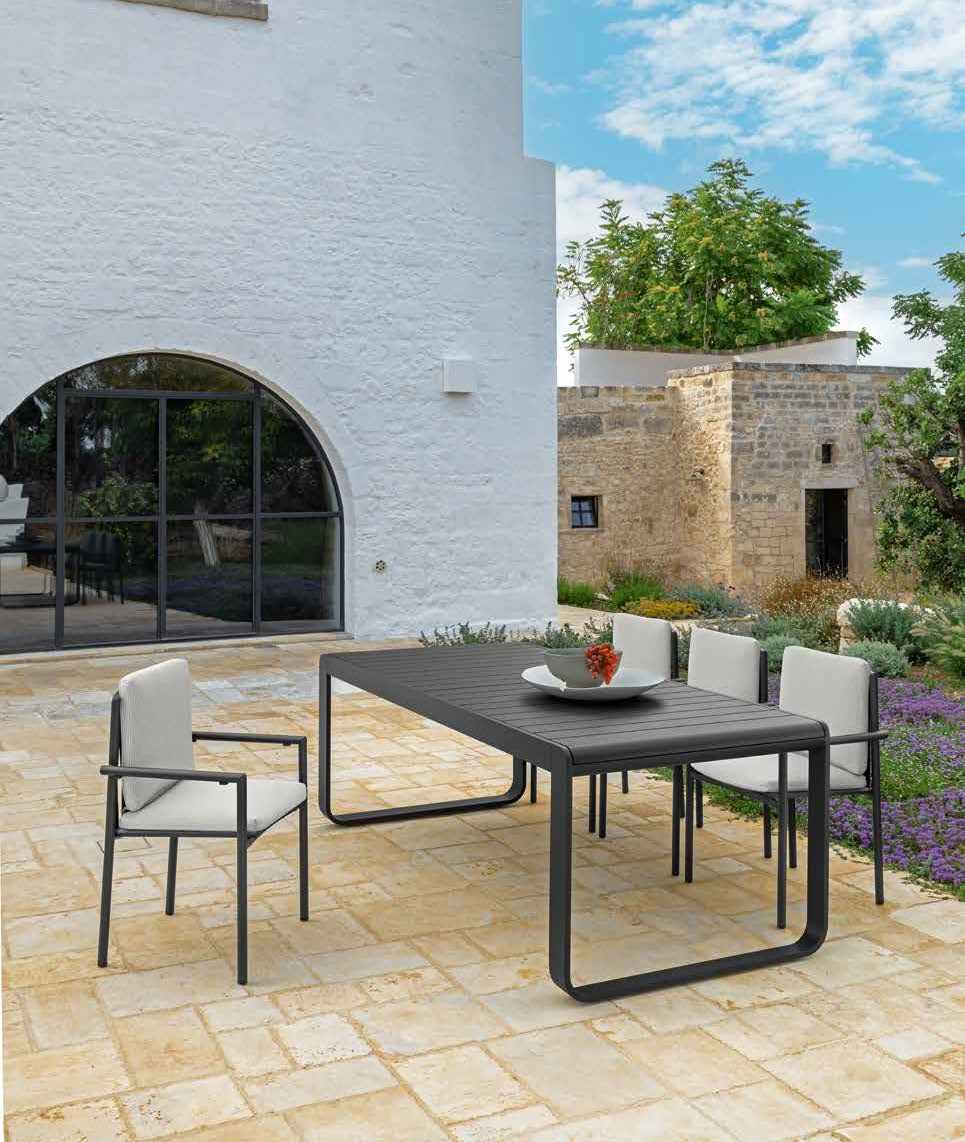 Un ensemble moderne de salle à manger d'extérieur composé de cinq chaises rembourrées et d'une table rectangulaire met en valeur l'aménagement extérieur d'un patio en pierre à côté d'un bâtiment blanc avec des fenêtres cintrées et une verdure luxuriante en arrière-plan.