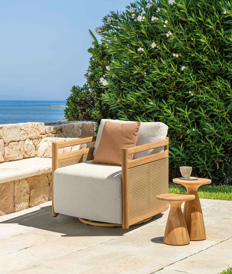 Un fauteuil en bois avec des coussins est posé sur un patio en pierre près d'une petite table d'appoint en bois, créant un aménagement extérieur confortable, avec de la verdure et l'océan visible en arrière-plan.