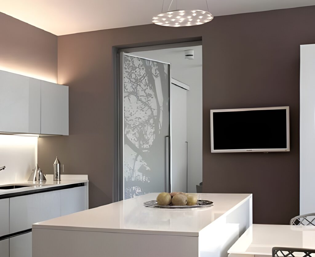 Cuisine moderne avec des armoires blanches, un îlot central surmonté de pommes, un téléviseur mural et une porte en verre dépoli ornée d'un motif d'arbre, mettant en valeur un aménagement intérieur élégant dans une palette de couleurs neutres et éclairé par un luminaire suspendu.